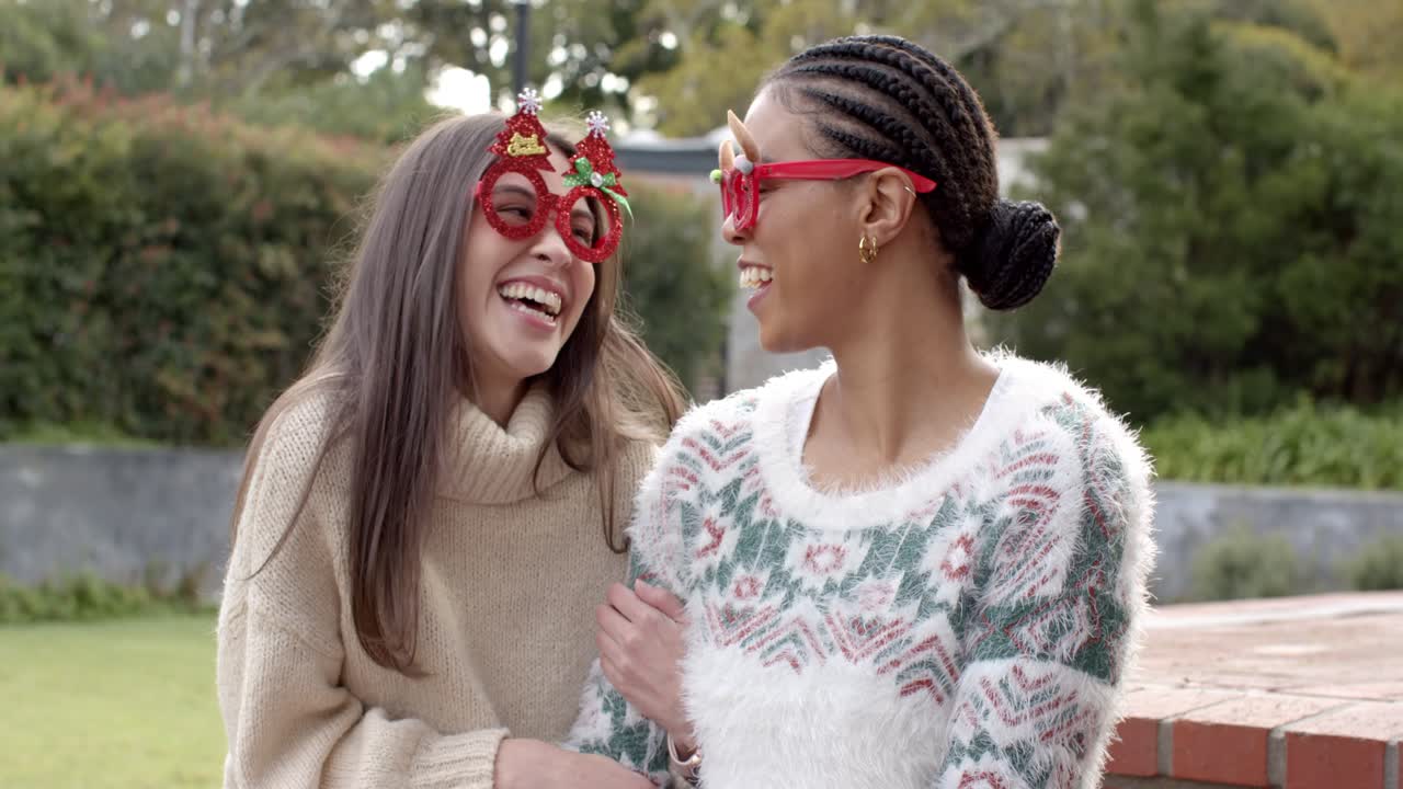Donning novelty glasses, diverse female friends wrapping arms and giggling in backyard