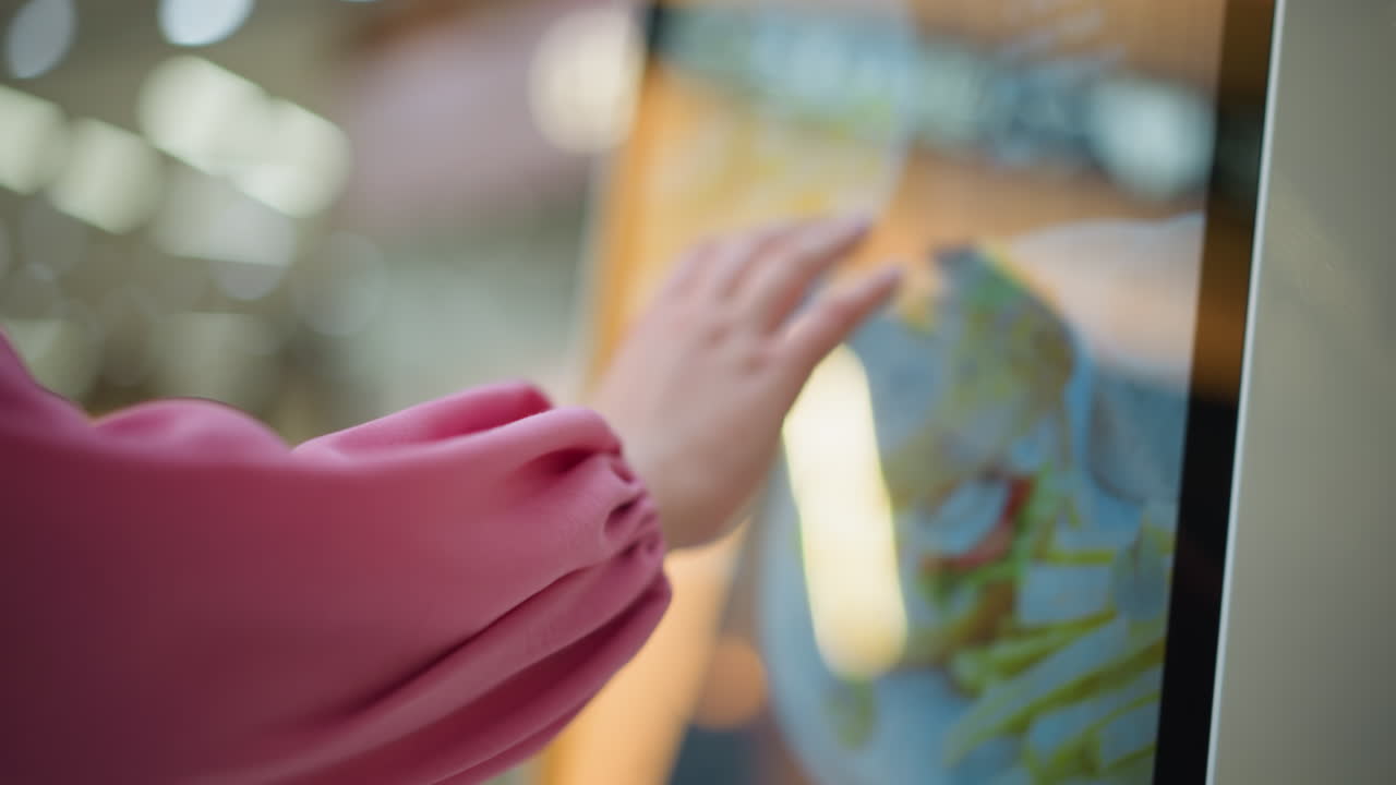 핑크색 셔츠를 입은 여성이 잘 조명 된 쇼핑몰에서 터치 스크린 기계와 상호 작용하는 손으로 볼 때, 화면은 변경 된 콘텐츠를 표시하지만 다른 항목과 배경 요소는 약간 흐릿합니다.