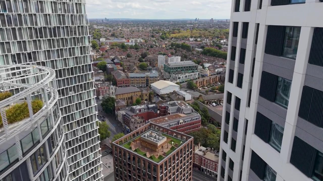 Il grattacielo Stratosphere e lo Stratford Eye visti durante un sorvolo in avanti con drone, che si muove tra i moderni grattacieli sopra le strade e i tetti di Stratford sotto cieli azzurri nuvolosi.