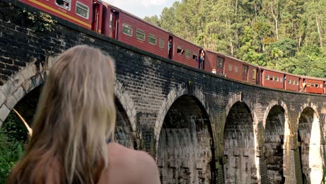 a young woman stands at a viewpoint near the nine arch bridge in ella, sri lanka, gazing at a colorful train as it moves across the historic stone arches.