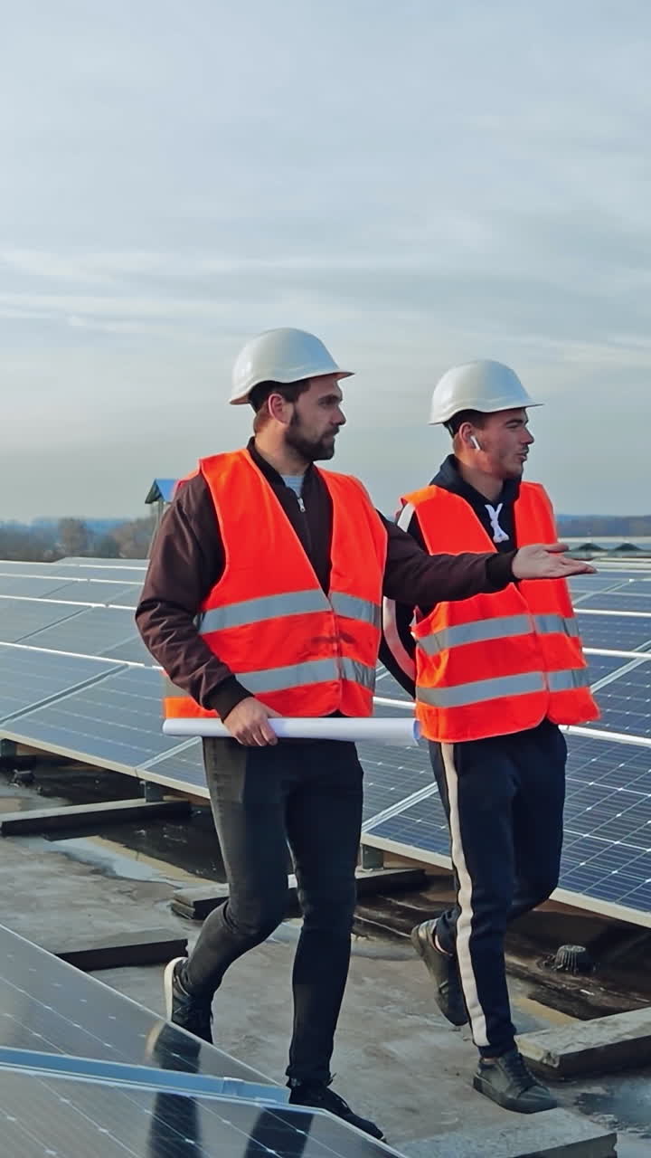 Installation of solar panels. Two workers in protective uniform and hard hats walking along the solar panels on the new photovoltaic station. Vertical video