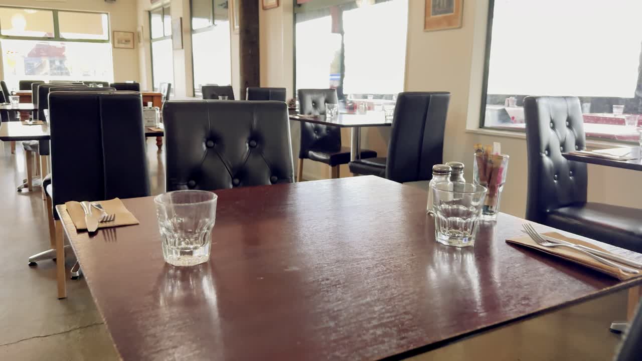 Camera slowly pans across a sunlit, modern cafe interior with empty tables, black chairs, glassware, and cutlery, emphasizing a calm, inviting atmosphere