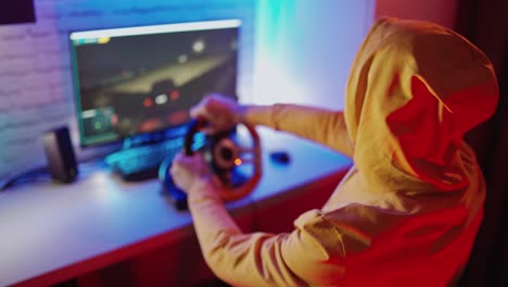 gamer in orange hood playing video game. young man enjoys playing car racing with steering wheel at home. entertainment concept.