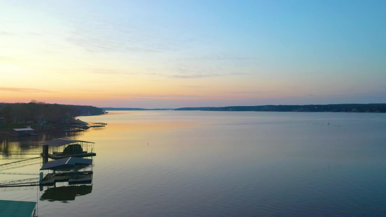 Večerní letecký pohled na klidné obytné jezero s zalesněnými kopci, domy u jezera, soukromými moly, tichými zátokami a pastelovým světlem západu slunce třpytícím se na otevřené vodě za letního soumraku.