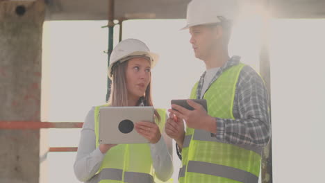 Ingenieure-Und-Designer,-Ein-Mann-Und-Eine-Frau-Stehen-Auf-Dem-Dach-Eines-Im-Bau-Befindlichen-Gebäudes-Und-Besprechen-Den-Plan-Und-Den-Baufortschritt-Mithilfe-Eines-Tablets-Und-Eines-Mobiltelefons.-Moderne-Bauarbeiter-Besprechen-Die-Infrastruktur-Des-Gebäudes-Und-Der-Umgebung