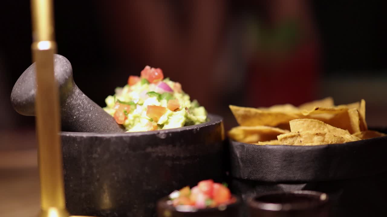 Close-up of guacamole and chips in a dimly lit Bangkok restaurant. A hand reaches for chips, creating a lively dining atmosphere