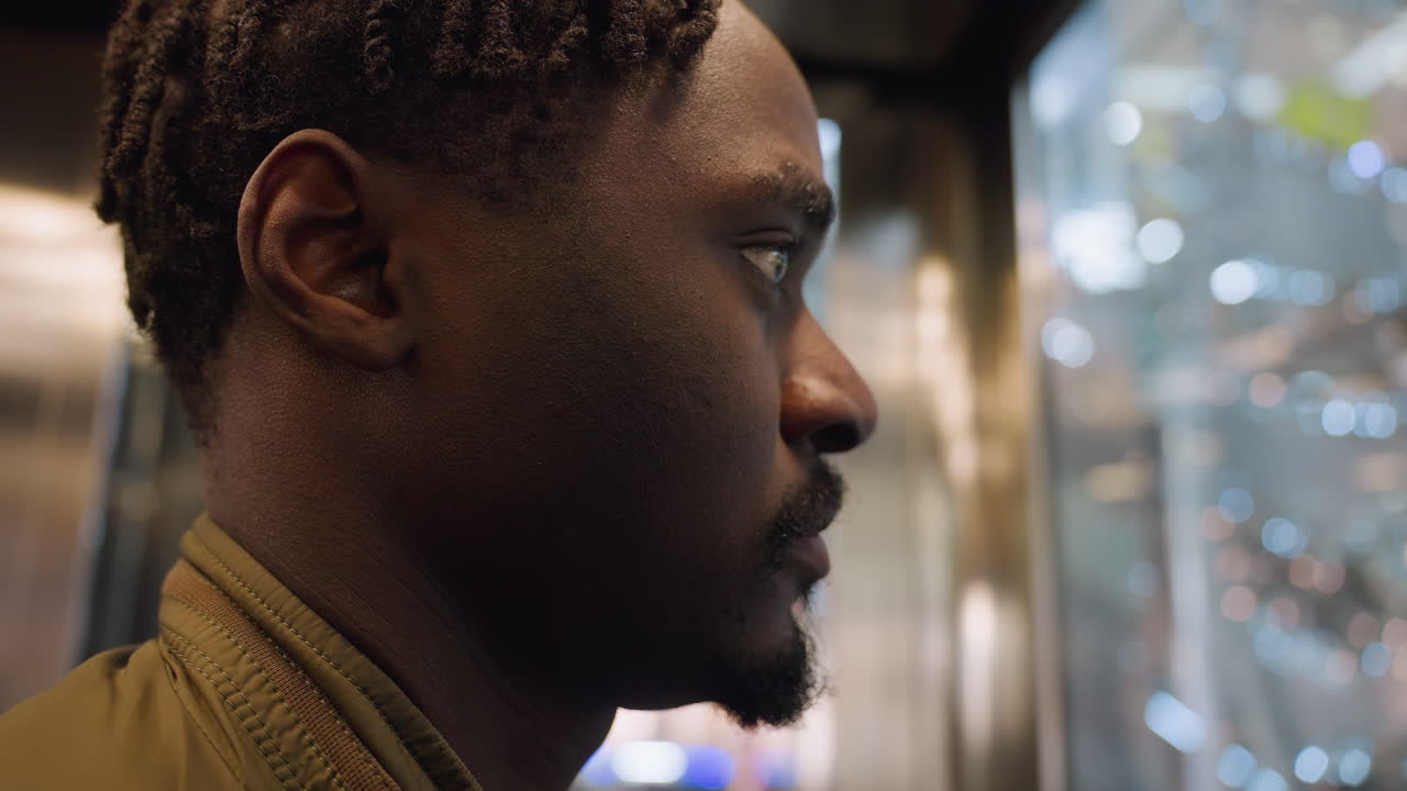 Close up of young man with braided hairstyle and trimmed beard wearing casual jacket standing inside moving elevator, gazing thoughtfully out of glass wall as city lights