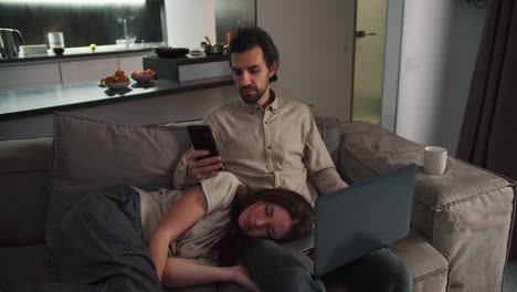 Serious-brunette-man-with-stubble-in-a-beige-shirt-works-on-a-black-phone-and-a-gray-laptop-while-sitting-on-the-sofa-while-his-happy-brunette-wife-lies-on-his-lap-in-a-modern-studio-apartment-in-the-evening