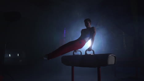 male gymnast athlete performs handstand and spin on pommel horse on a dark background and smoke in slow motion. olympic programme. preparation for the exercise