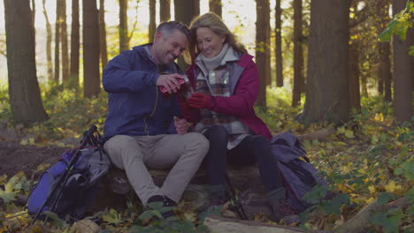 mature couple sitting on log stop for rest and pour hot drink from flask on walk through fall or winter countryside - shot in slow motion