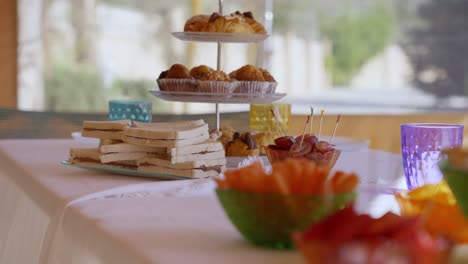 Mesa-Con-Dulces-Para-Una-Fiesta-En-Verano,-Con-Una-Bandeja-Con-Croissants,-Sandwich,-Crudités,-Vasos-De-Colores-Y-Mantel-Blanco-Moviéndose-Por-El-Viento