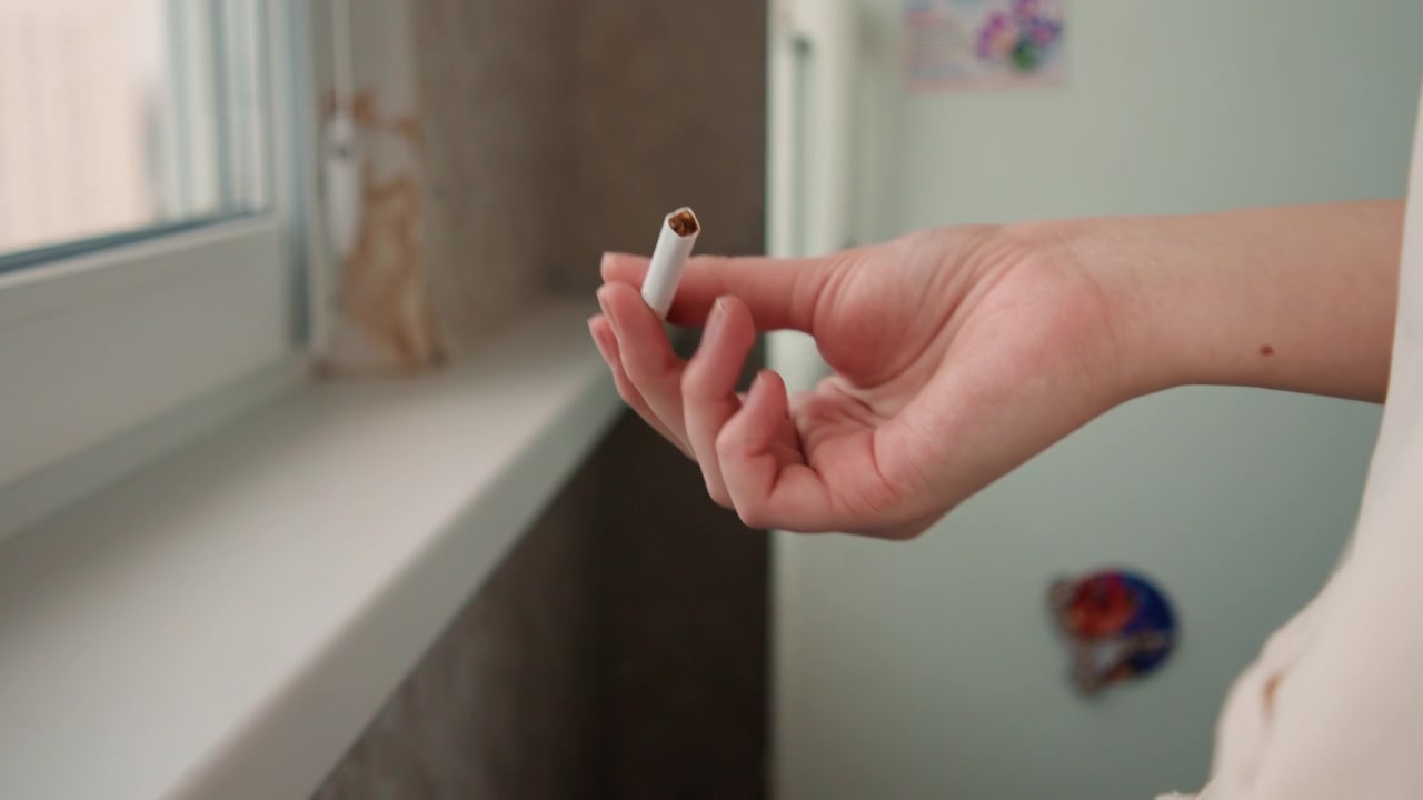 Una persona hace una pausa con un cigarrillo en la mano. Manos sujetando un cigarrillo terminado junto a la ventana en un momento de reflexión. Primer plano de manos sosteniendo un cigarrillo completado en el alféizar de la ventana durante una tranquila tarde urbana.