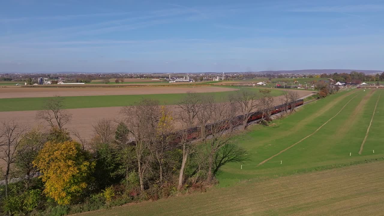 Una vista aerea di un singolo binario ferroviario che attraversa paesaggi agricoli di campagna mentre un treno a vapore si avvicina in lontananza in una soleggiata giornata autunnale.