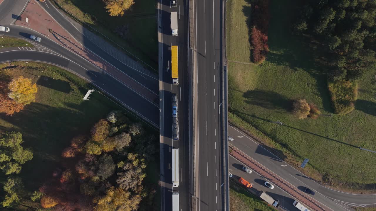 Top-down aerial view of a highway showing several trucks and cars moving in both directions. The surrounding landscape features green fields and autumn trees, illustrating modern road transport