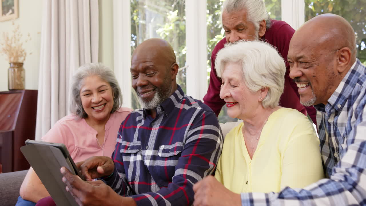 Amigos idosos diversos e felizes, homens e mulheres, usando tablet juntos em uma sala de estar ensolarada, em câmera lenta.