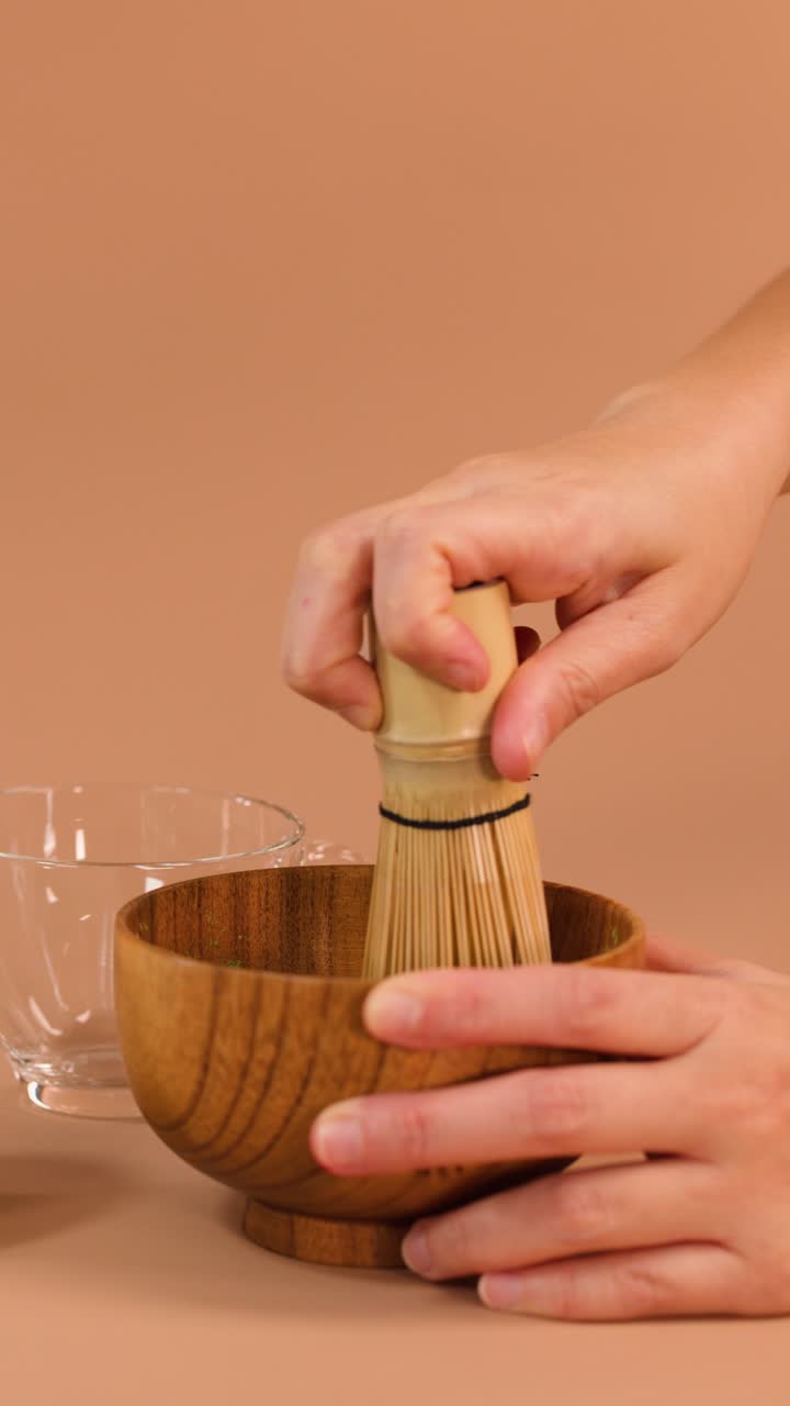 Mano che usa una frusta di bambù per mescolare la polvere di matcha in una ciotola di legno, sfondo neutro, illuminazione soffusa.