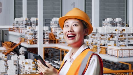 vista lateral de cerca de una ingeniera asiática con casco de seguridad usando un teléfono inteligente mientras está de pie en el almacén con estantes llenos de productos de entrega