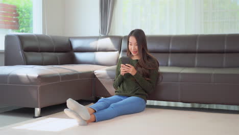 asian woman sitting on the floor near sofa texting on smartphone