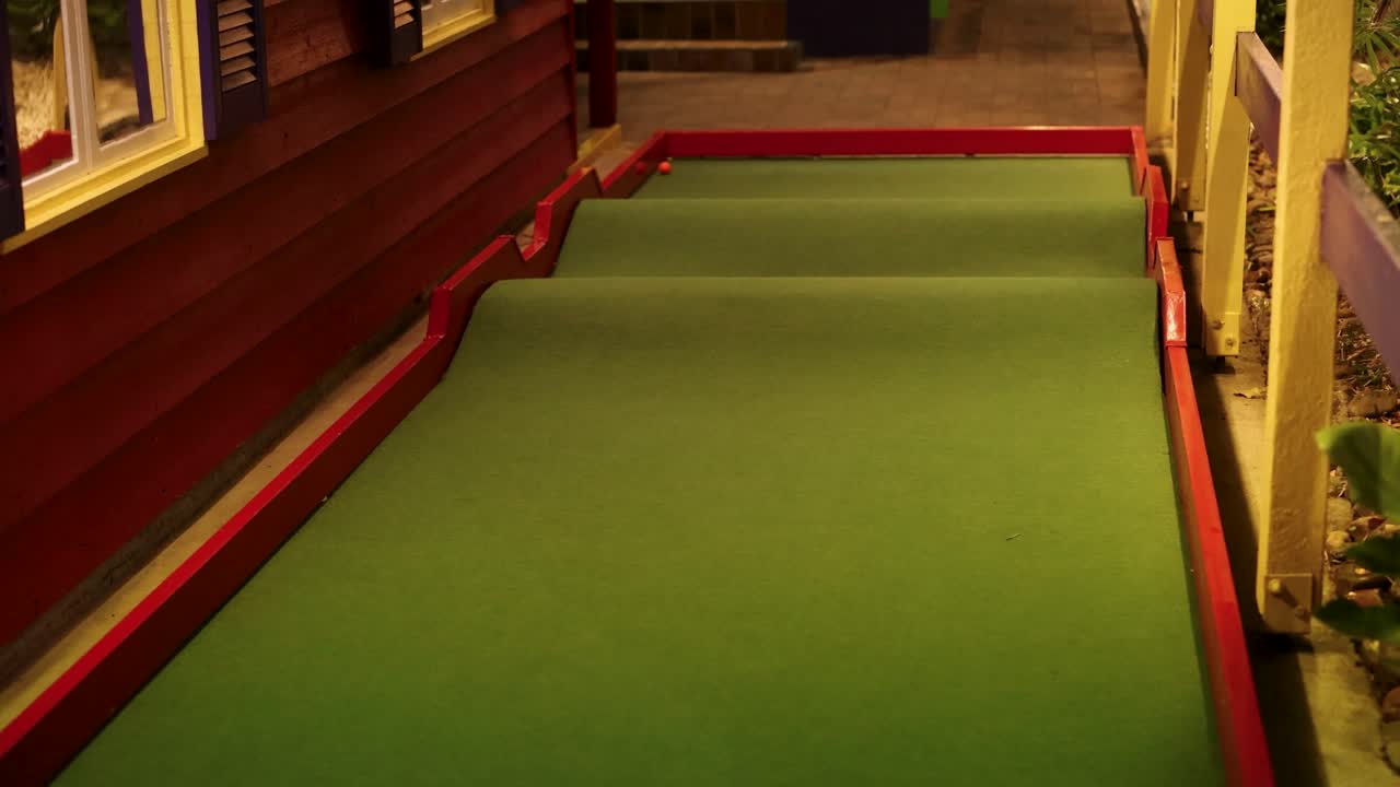 una pelota de golf rueda a lo largo de un campo en miniatura por la noche, capturada en una iluminación cálida con ángulos de cámara estables