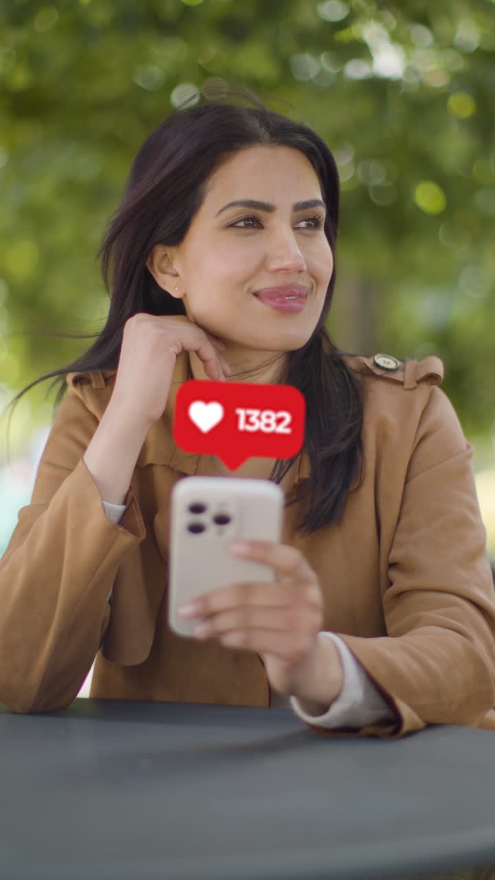 Vertical Video Of Smiling Woman Sitting At Outdoor Table Looking At Mobile Phone With Motion Graphics Emojis Showing Multiple Social Media Notifications Liking And Reacting To Online Content