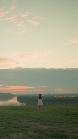 vista trasera de una mujer con un vestido blanco y sombrero de pie sola en un campo de hierba aislado por un lago tranquilo al atardecer. ella mira el entorno sereno, abrazando el momento pacífico de la soledad