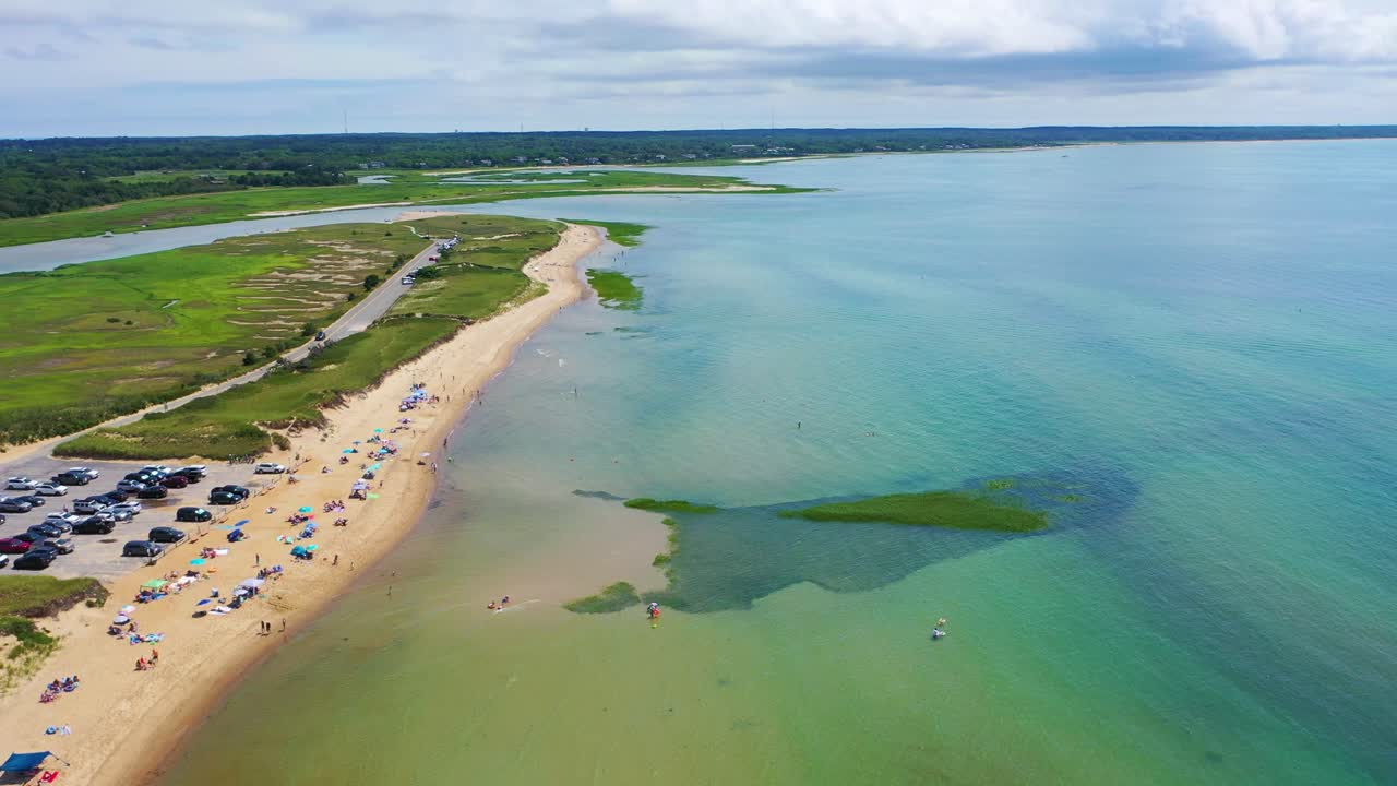 Drone footage captures a vibrant shoreline with beach houses, parked cars, families under umbrellas, and swimmers enjoying turquoise waters, while a small boat floats just offshore near the coast
