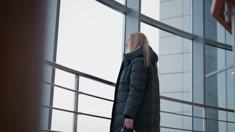 young lady walking near iron railing, gliding hand along metal rail, with city view through glass windows, wearing stylish winter coat, handbag in hand, reflecting urban life around