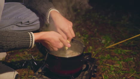 close up of person hands peeling potatoes with knife over steaming pot on portable stove during night camping in forest, surrounded by earthy ground and gentle warm light