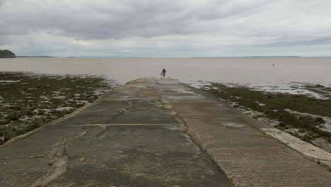 grandparent and grandchild explore coastal slipway edge watching the sea on cloudy day, wide shot