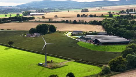 aerial view of a wind turbine slowly rotating above vibrant green and brown farmland near kinross, scotland, under bright daylight with soft shadows