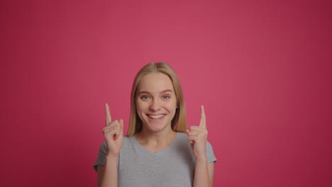 funny blonde girl smiling at camera and pointing at copy space over pink wall