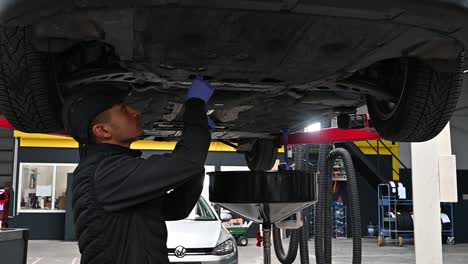 Young-mechanic-working-on-bottom-of-lifted-vehicle-inside-modern-garage,-slow-motion