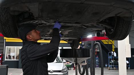 joven mecánico trabajando en la parte inferior de un vehículo levantado dentro de un garaje moderno, a cámara lenta