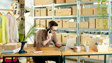 Caucasian-designer-man--in-black-mask-sitting-at-table-and-talking-on-smartphone-while-browsing-online-on-laptop-in-clothing-shop-warehouse