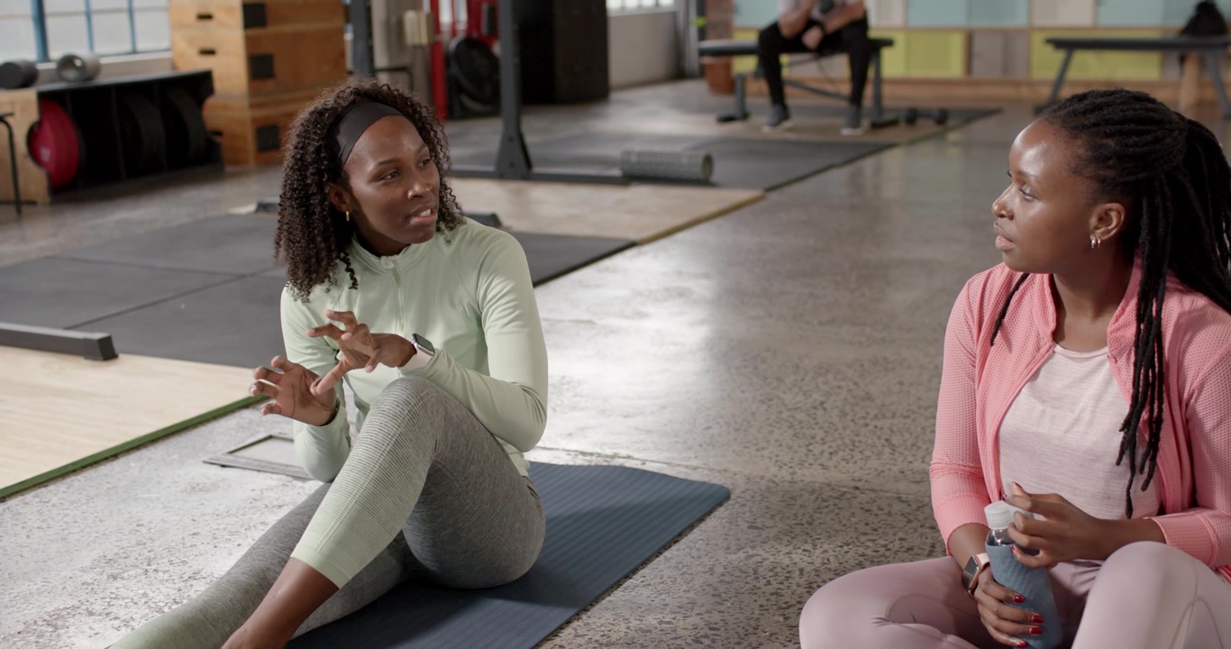African American women resting on mats at gym, left looking then talking, right holding blue bottle