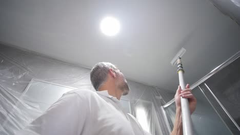 Man-In-White-Attire-Painting-Ceiling-With-Paint-Roller-On-Stick-In-Bathroom-Covered-In-Plastic-In-Slow-Motion