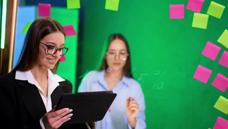 two workers in action. one woman in glasses uses a tablet while another observes in a bright, colorful office space during the day