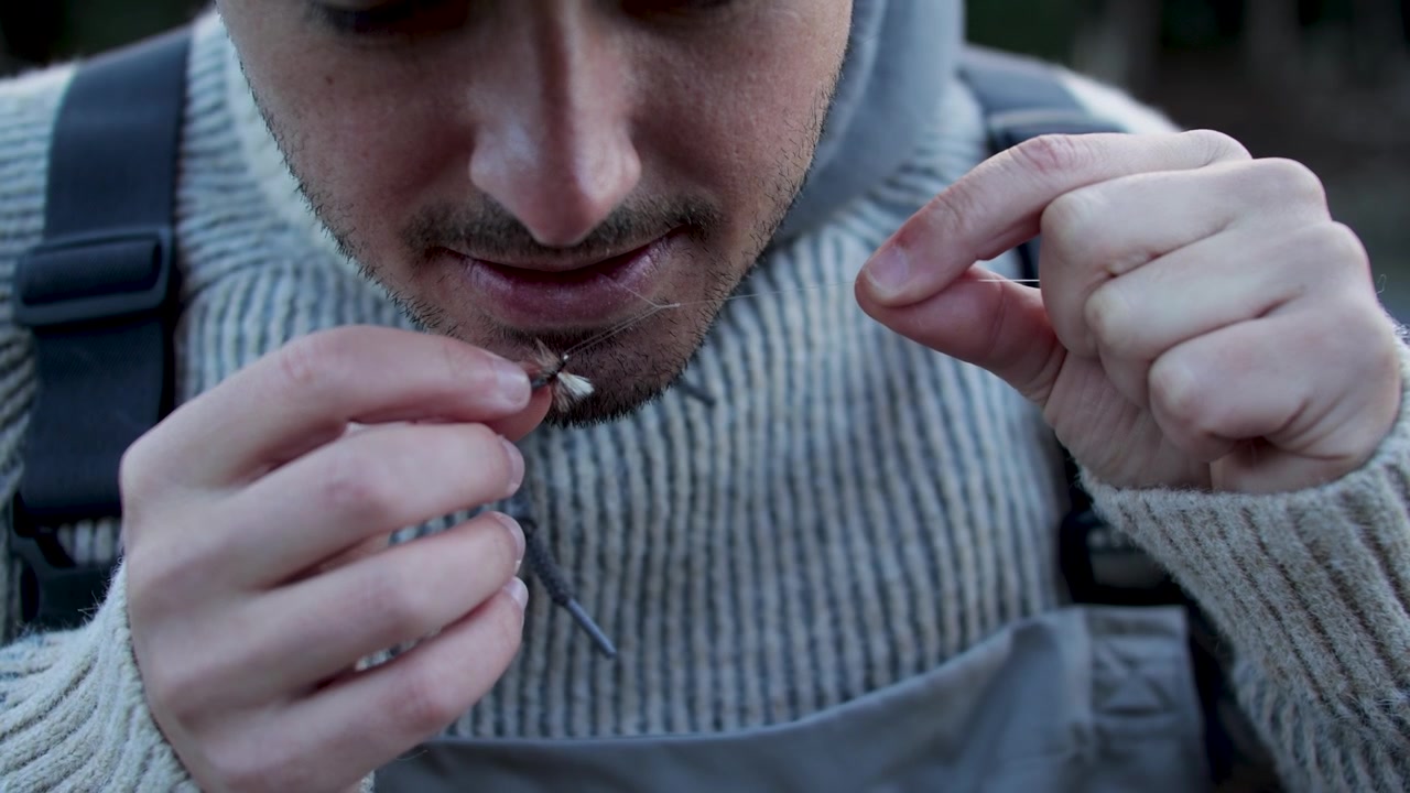 Primo piano fisso da terra di un pescatore che lega una mosca fresca alla sua canna da pesca a mosca accanto a un tranquillo lago alpino sulla Grand Mesa del Colorado, preparandosi a pescare trote iridee e salmerini di fontana.