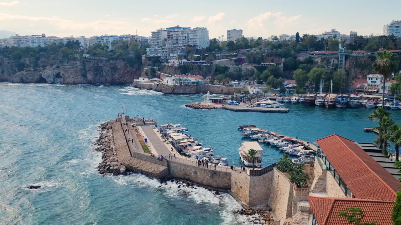 Windy spring day at the marina in Antalya, Turkey. Waves crash against the pier as pedestrians and tourists stroll along the waterfront promenade