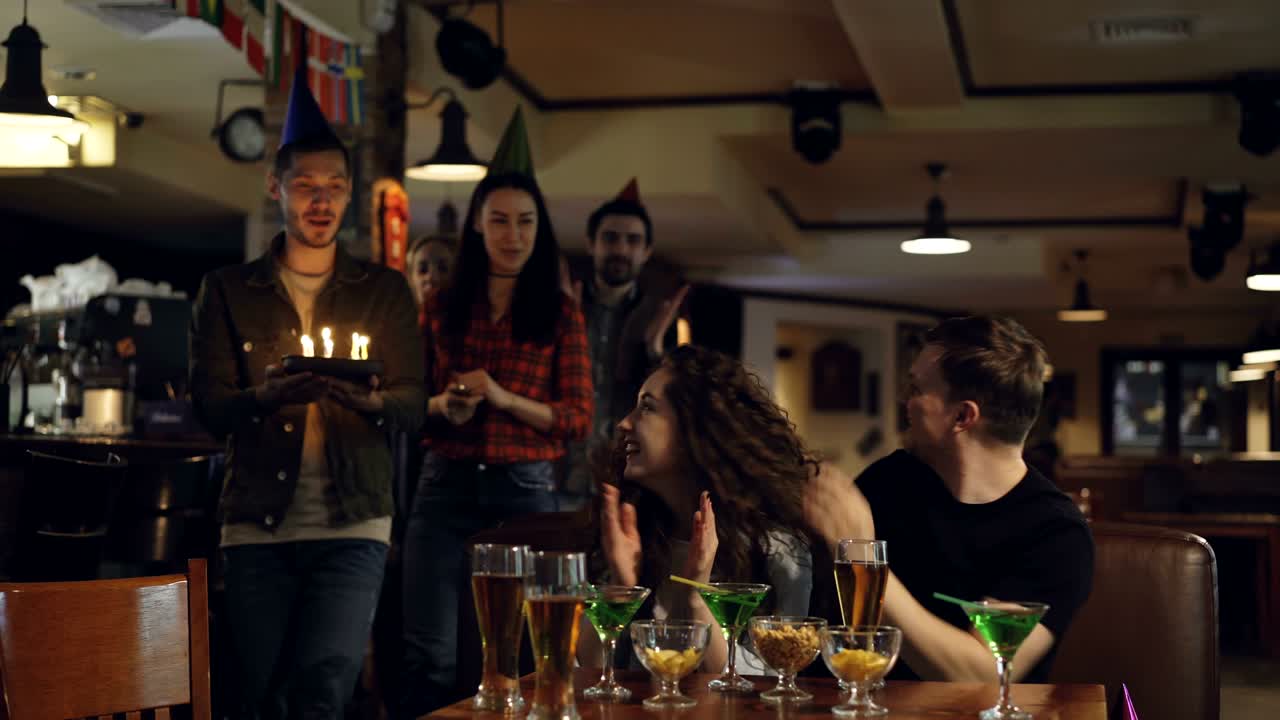 los amigos están trayendo pastel de cumpleaños a la bonita morena sentada en la mesa en el café con los ojos cerrados. ella está haciendo deseos y soplando velas, los amigos están aplaudiendo.