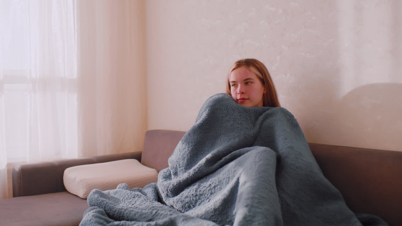 Woman sitting alone on sofa wrapped in fluffy blanket with closed eyes and calm face in soft morning light, expressing solitude and emotional stillness in peaceful cozy room near curtain and sunlight