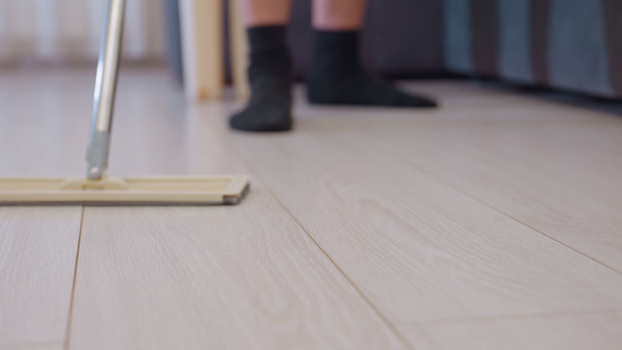 Lower angle close up of adult wearing black socks pushing mop across wooden floor near sofa during household cleaning task showing daily domestic routine focused on hygiene