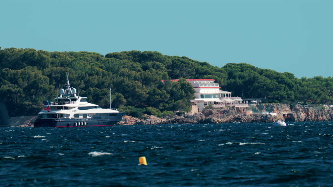 A luxurious white yacht anchored near a rocky Mediterranean coastline surrounded by lush green pine trees and elegant seaside villas