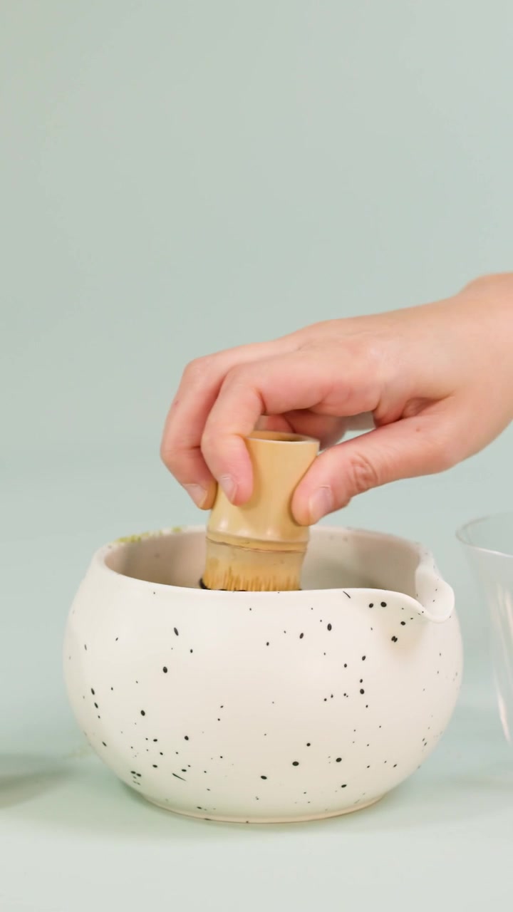 Una mano usa un frullino di bambù per preparare il tè matcha in una ciotola bianca maculata