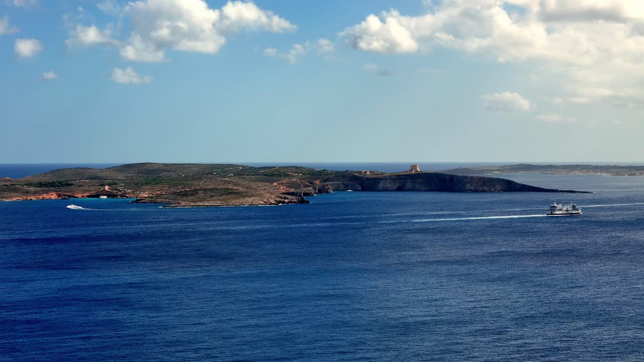 A wide angle view of the historical Saint Mary's Tower on Comino Island under a clear summer sky during a journey from Malta to Gozo across the crystal turquoise Mediterranean sea with ferry passing
