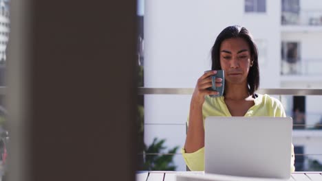 Mixed-race-gender-fluid-person-drinking-cup-of-coffee-and-using-laptop-on-roof-terrace