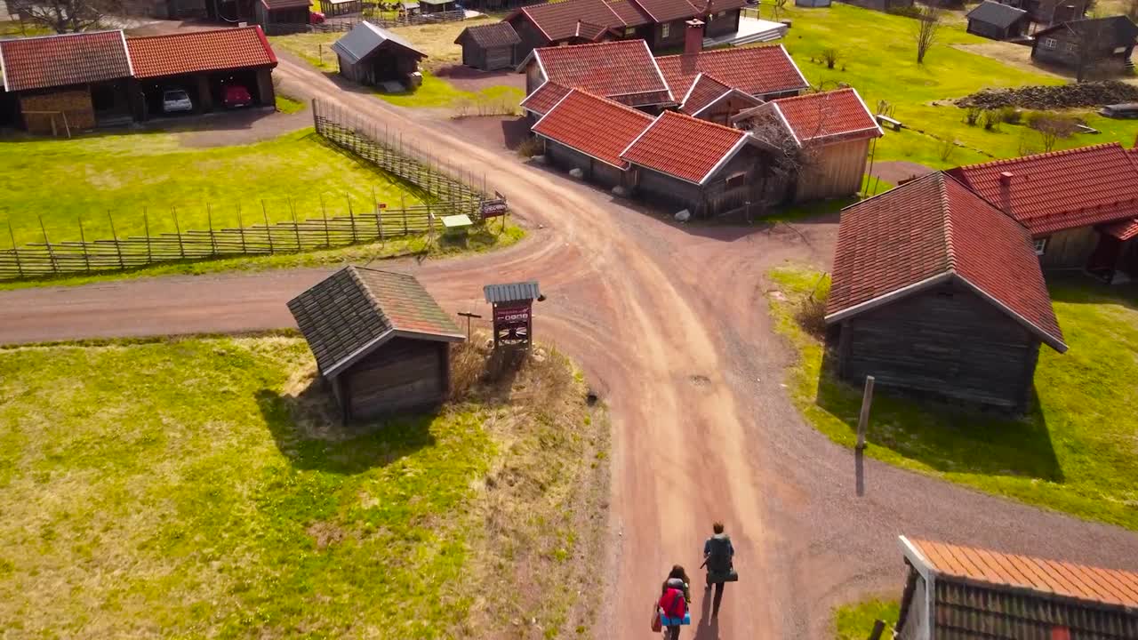 Aerial drone footage of a couple of backpackers or hiker walking on a muddy road between cute little houses or villages in Sweden that have red rooftops and are surrounded by green grassy lawns, sunny