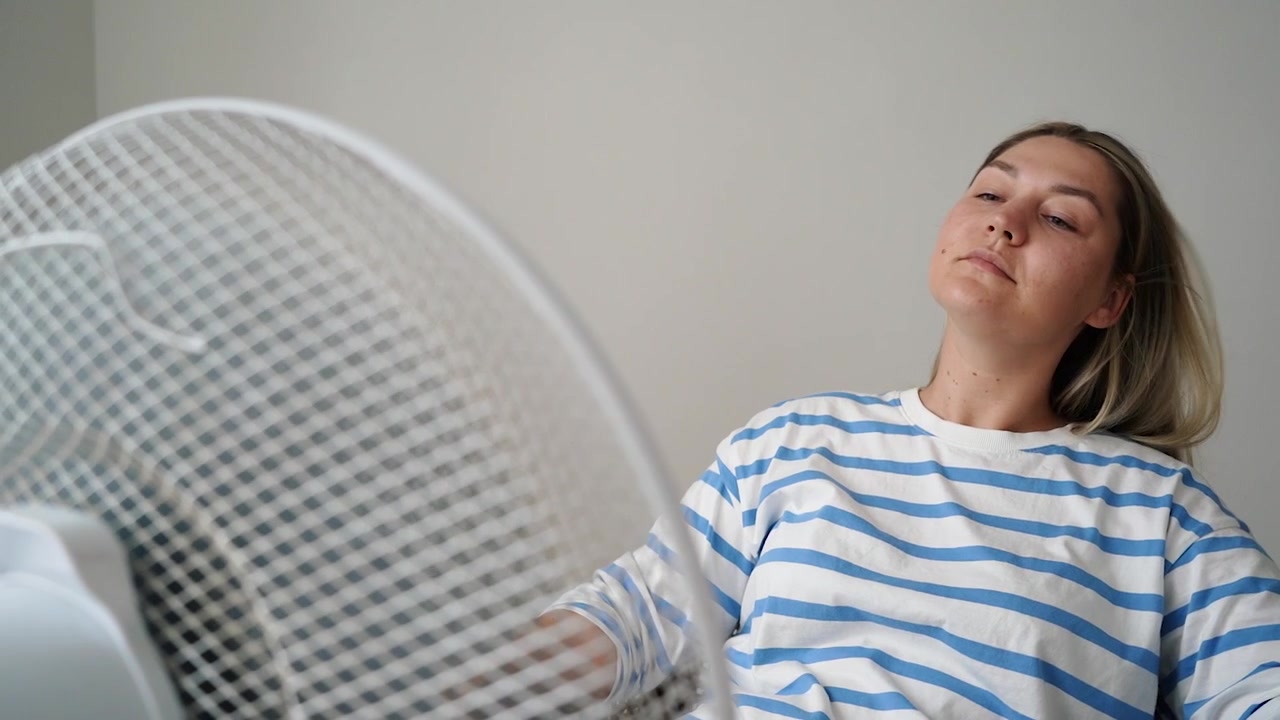 Mujer acalorada con camiseta de rayas sentada frente a un ventilador doméstico, con los ojos cerrados y el cabello al viento mientras se refresca.