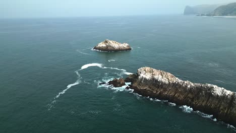drone flies backward over ocean, revealing rocky islands with birds off the coast of lima, peru. soft morning light with calm sea and clear sky.