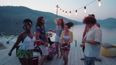 young friends dancing with drinks at lake party on pier